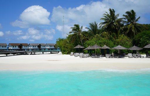 turquoise water laps a white beach with thatched umbrellas and huts and palm trees