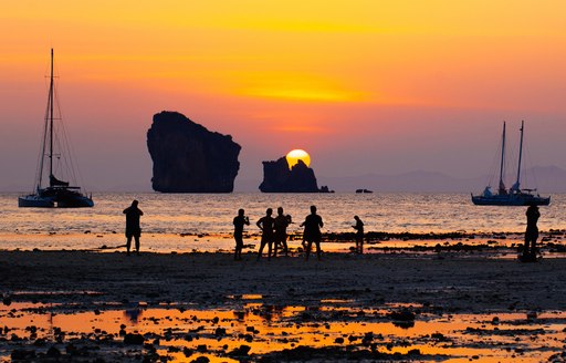 a sun sets behind some rocks on a bay in thailand with yachts and fishermen silhouetted