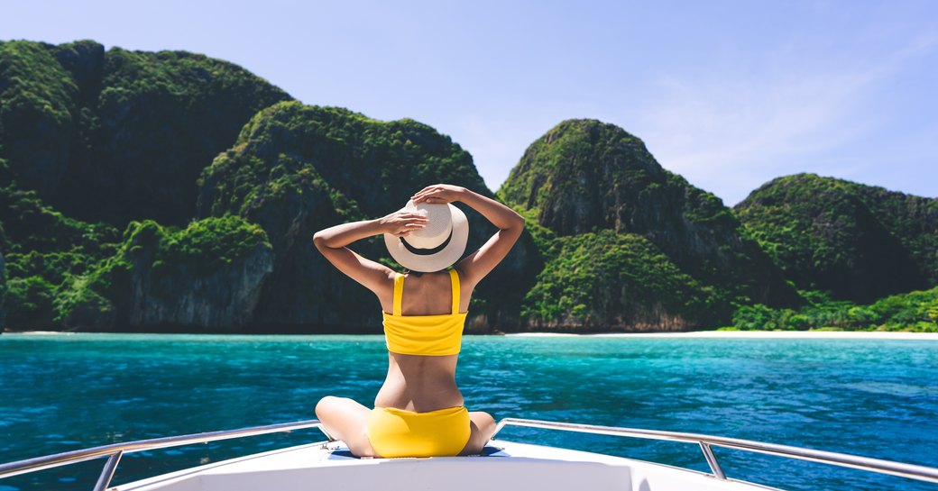 Woman in a yellow bikini on the bow of a yacht looks out towards lush hilly emerald island in Thailand